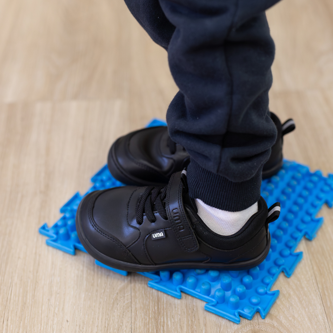 Zapatilla escolar barefoot negra modelo Dash con doble velcro para kids en uso sobre superficie sensorial
