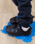 Zapatilla escolar barefoot negra modelo Dash con doble velcro para kids en uso sobre superficie sensorial
